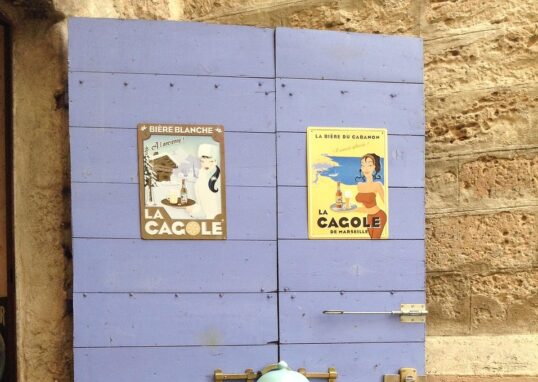 Vieille porte en bois de couleur lavande avec deux affiches vintage avec le mot CAGOLE et une fontaine en émail clouée sur la porte avec savon de Marseille