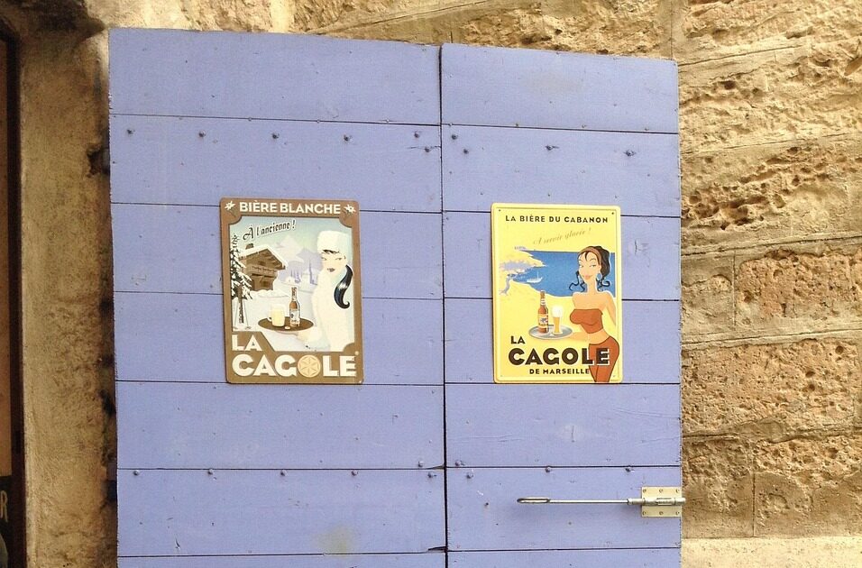 Vieille porte en bois de couleur lavande avec deux affiches vintage avec le mot CAGOLE et une fontaine en émail clouée sur la porte avec savon de Marseille