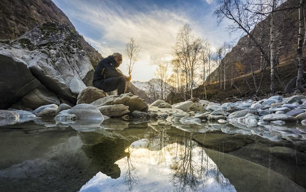 aua fond d'une vallée de montagne en hiver un homme esta ssis sur des rochers au borsd d'une rivière et regarde l'eau il semble tenir une branche dans se mains c'est en fait une illusion car il s'agit d'un arbre en second plan