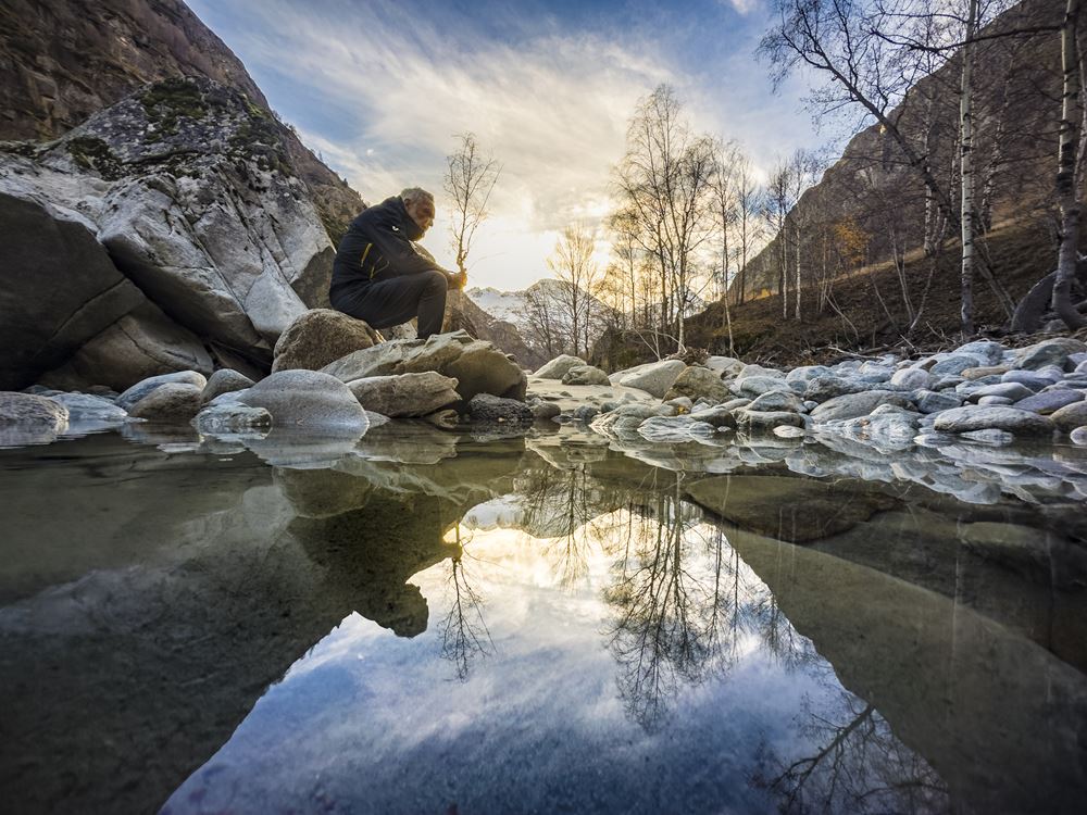 aua fond d'une vallée de montagne en hiver un homme esta ssis sur des rochers au borsd d'une rivière et regarde l'eau il semble tenir une branche dans se mains c'est en fait une illusion car il s'agit d'un arbre en second plan